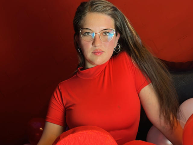 AuroraCou with long brown hair, glasses, red turtleneck, and hoop earrings, sitting, holding a red pillow, looking at the camera, red background, soft lighting, slightly bluish reflection.