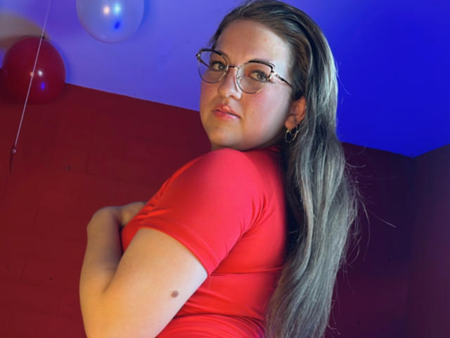 AuroraCou with long brown hair, glasses, and red shirt, looking back, holding her tits with her arm. Blue and red wall, balloons in background.