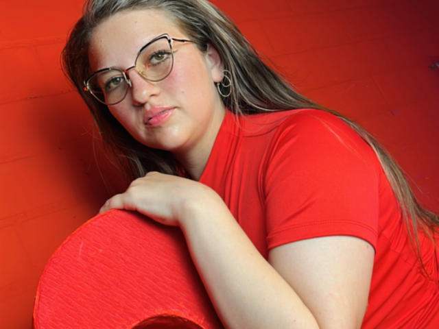 AuroraCou with glasses, long brown hair, red shirt, holding a red cushion, looking slightly annoyed, wearing hoop earrings, in a red-walled setting.