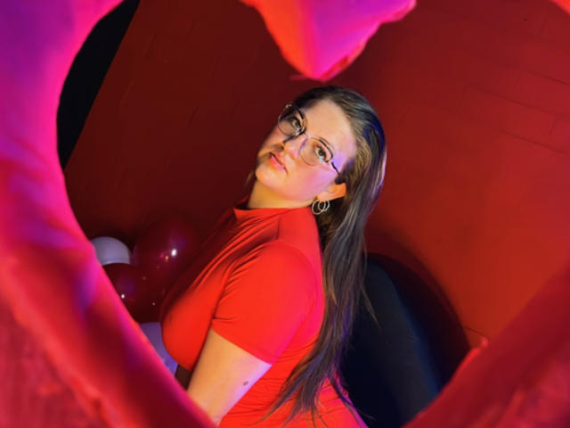 AuroraCou with glasses, red shirt, and earrings, looking back, her long brown hair, small tits, and red balloon in foreground, pink and red lighting, confident expression.