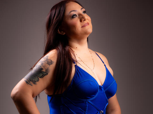LadyNylonOh with dark red hair, blue bra, and butterfly tattoo on her right arm, wearing a gold necklace, smiling slightly, looking up, hands on hips, studio light.