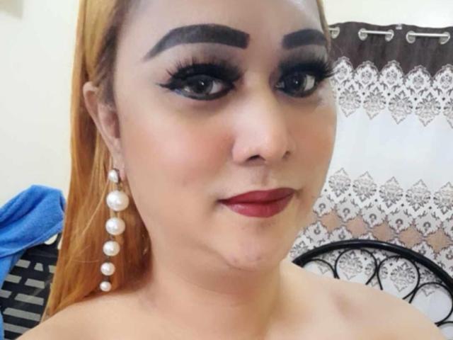 BigBoobsBigcum has thick, dark eyebrows, long eyelashes, red lipstick, and pearl earrings. She's smiling with a small scar under her chin, and has orange hair.