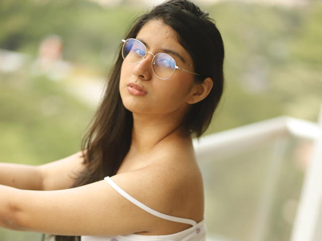 TereCarpert with long dark hair, wearing round glasses and a white strap top, showing her shoulders and upper chest, looking slightly to the side. Soft, natural light.