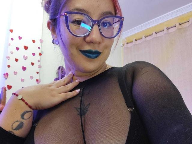 HollyManziniBBW with blue lipstick, glasses, and black sheer top, showing a tattoo on her chest and a hand with a red bracelet, smiling at the camera.