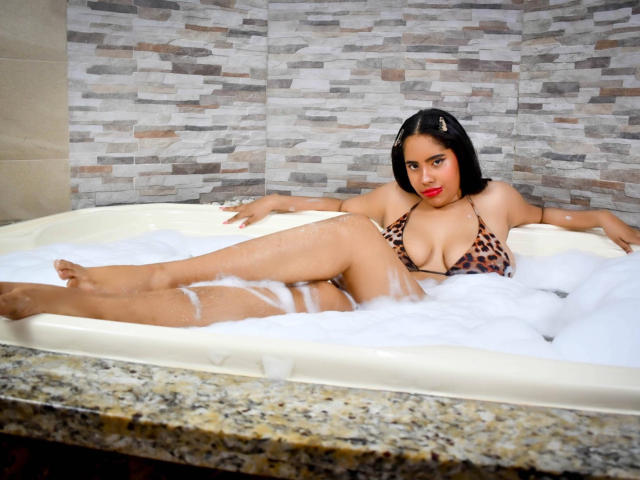 SiennaTaylor is in a bathtub with white foam, wearing a leopard print bikini, her ass and tits partially visible, with black hair and red lipstick, looking at the camera.
