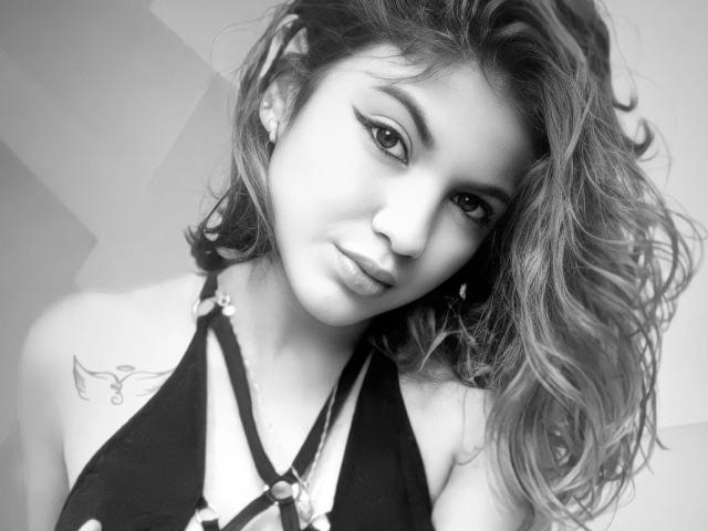 EmyBecker has wavy hair, a tattoo on her left shoulder, and wears a black strappy top, looking down with a slight smirk. Black-and-white photo, focus on face.