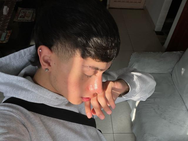 ImJuanda with a shiny nose, black ear piercing, short dark hair, gray hoodie, and black straps, eyes closed, hand near face, on a light gray couch.