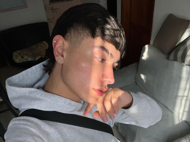 ImJuanda, with short black hair, a nose ring, and a silver earring, leans on a gray couch in sunlight, wearing a gray hoodie, with a black strap.