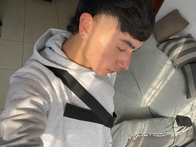ImJuanda with short dark hair, a nose ring, and ear studs, wearing a gray hoodie and black seatbelt, sitting on a light green couch, eyes down, in sunlight.