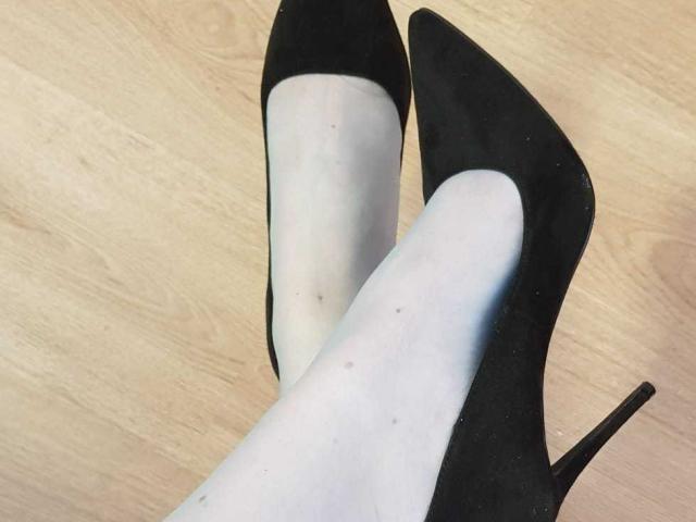 Las piernas de MilfXX, con tacones altos negros de punta y tacón de aguja, llevando medias blancas transparentes, están sobre un suelo de madera. Sus tacones están ligeramente cruzados..