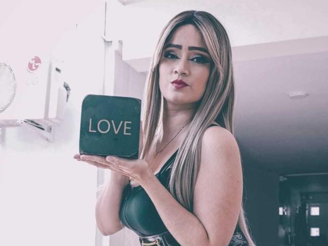 EmmaGoldyx with long blonde hair, red lipstick, black leather top, holding black box with "LOVE" in gold letters, pouting, looking at camera.