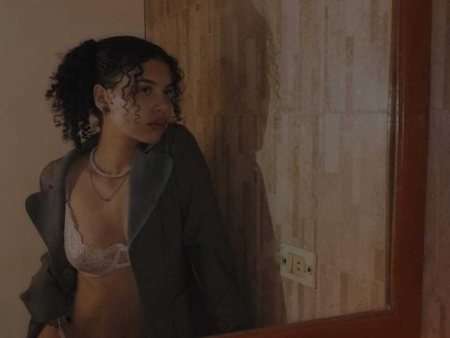 NataliArt with curly black hair, wearing a sheer green shirt and white lace bra, standing in a dimly lit room, looking to the side with a subtle, confident smile.