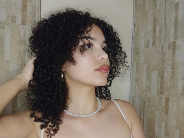 NataliArt, met krullend zwart haar, draagt een witte top met bandjes en een parelketting, en kijkt zijwaarts met een hand in haar haar. Ze heeft roze lippenstift en een serieuze uitdrukking..