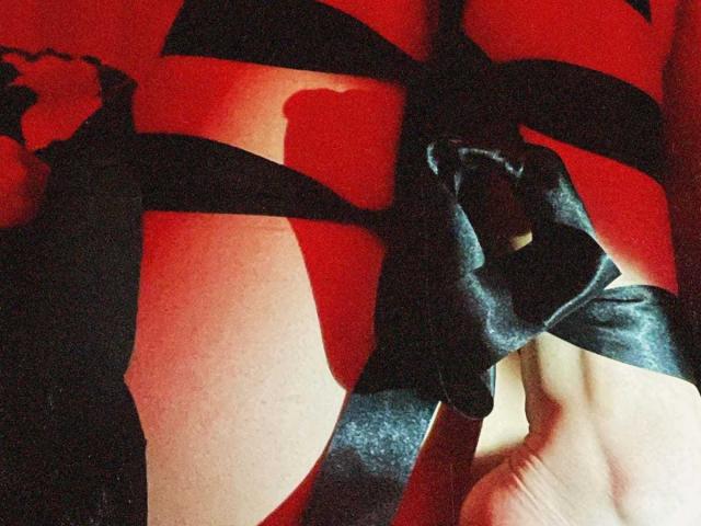 NataliArt's bare butt wrapped in black satin ribbon, showing shadowed creases and soft skin texture, with a hand gently holding the ribbon on the right side.