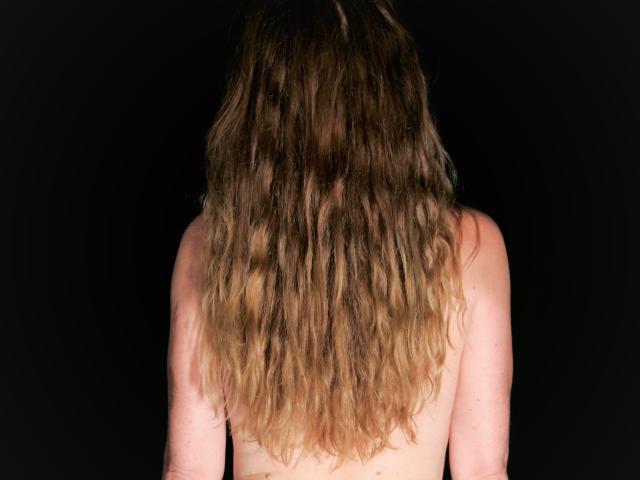 MilfXX regresa con su largo cabello ondulado castaño cubriendo sus hombros desnudos, sobre un fondo negro. Está sin sujetador, y su trasero es ligeramente visible en la parte inferior..