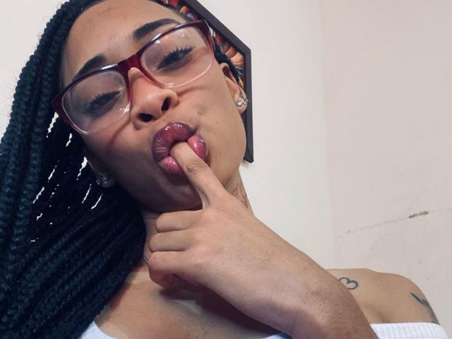 ElaHott with dark braids, red glasses, and pink lipstick, sucking her finger, showing her tongue. She's got a tattoo on her left shoulder and earrings in.