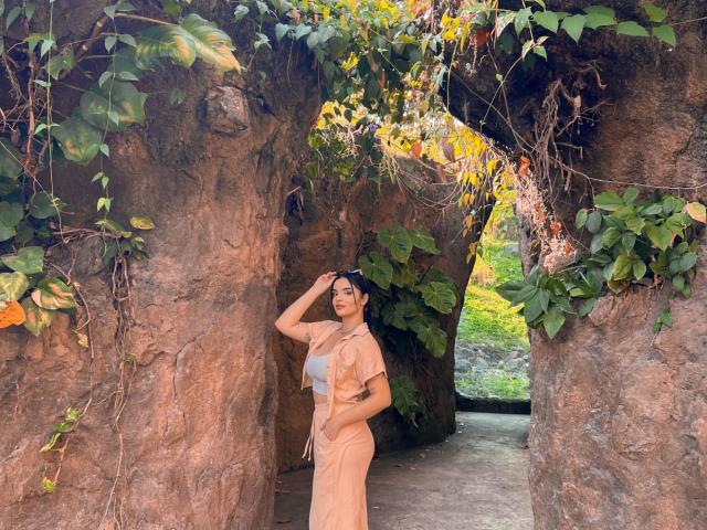 NovaDixon stands in a rocky, vine-covered archway, wearing a beige dress, with one hand on her head, showing her ass, and looking at the camera.