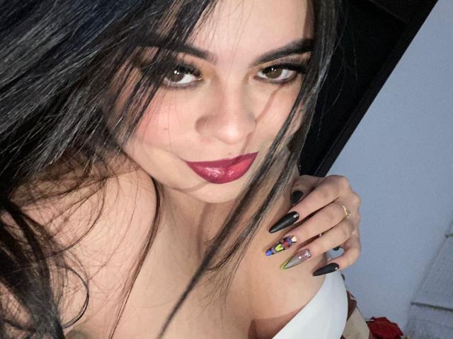 MeliPink with long black hair, dark eyes, and red lipstick, showing off colorful nails, smiling slightly, wearing a white top, and holding her shoulder. Boobs visible.