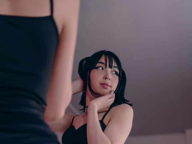 MillaHottLove69 with black hair, looking in mirror, wearing black dress, hand on neck, reflection shows her face, upper body, and another person's arm in foreground.