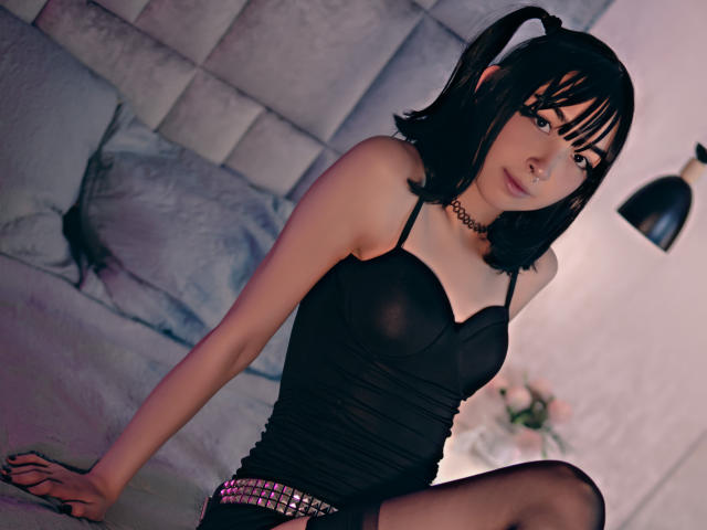 MillaHottLove69, con due codini neri, indossa una maglia nera trasparente, una collana e una cintura con borchie, seduta su un letto, mostrando i suoi piccoli e carini seni e un leggero, sexy sorriso..
