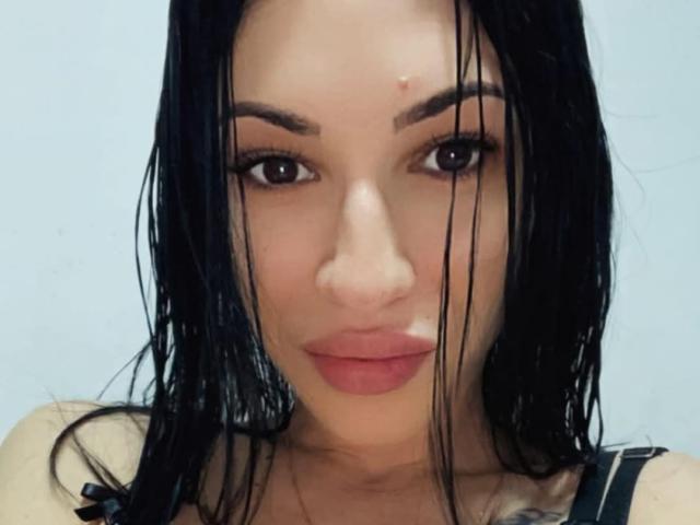 DenysaD has wet black hair, brown eyes, and a small red bump on her forehead. She's wearing pink lipstick and a black strap top, showing her tits.