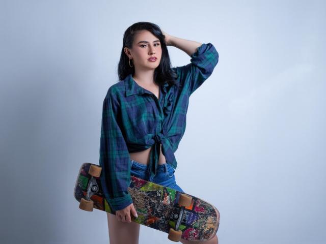 FrancheskaWaalsh with dark hair, green-blue plaid shirt tied at the front, blue shorts, holding a colorful skateboard, hand in hair, standing, looking at camera, slightly pouty lips.