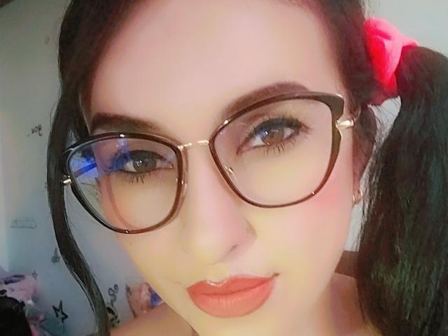 LuluFrancaise has fair skin, black pigtails with red scrunchies, and wears big black glasses. She has red lipstick and blue eyes, looking slightly up at the camera.