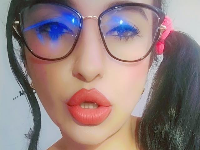 LuluFrancaise with blue-tinted glasses, pink scrunchie in black pigtails, red lipstick, and slightly open mouth showing teeth, looking up with reflection on glasses.
