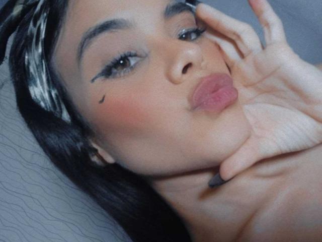 SophieAlessia69, met volgezwollen lippen, donker haar en zwarte eyeliner, die haar gezicht aanraakt met een hand, waarbij een kleine moedervlek op haar rechterwang zichtbaar is. Haar borsten zijn niet zichtbaar..