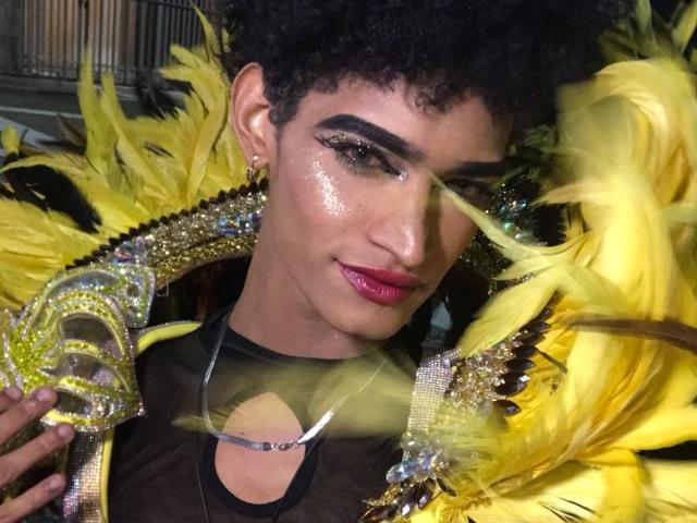 AdharaX69 with shiny skin, pink lipstick, and glitter, holding a yellow feathered mask with gems, wearing a black sheer top, and curly black hair. Boobs partially visible.