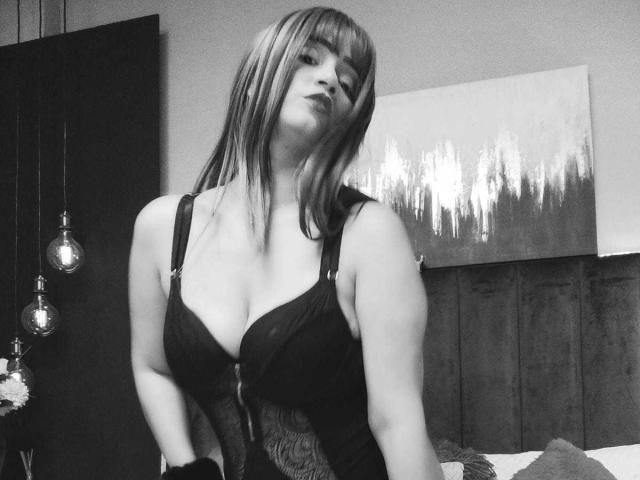 Black-and-white photo of EmmaGoldyx with straight bangs, wearing a lace-up black bra that highlights her large tits. She has a confident, slightly tilted head and a seductive look.