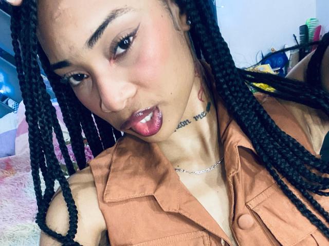 ElaHott with brown skin, black braided hair, and pink lips, wearing a tan button-up shirt, tongue slightly out, showing a tattoo on her neck, looking seductively at the camera.