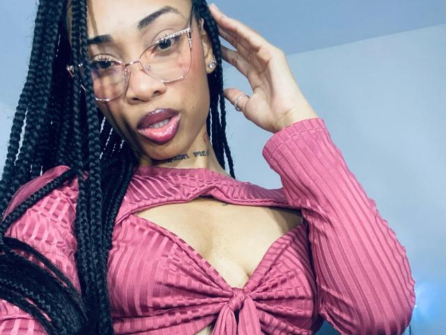 ElaHott, avec de longues tresses noires, des lunettes roses et un haut rose côtelé qui dévoile sa poitrine, une main dans les cheveux, du rouge à lèvres rose, un tatouage au cou, une peau claire, une expression confiante..