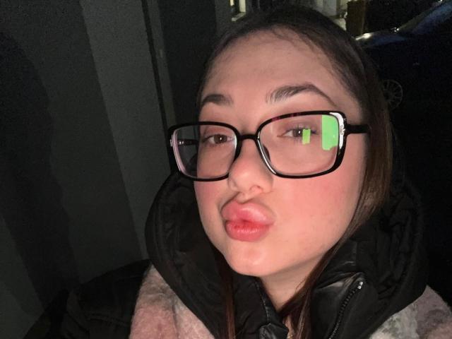 MayAlisa with puffy pink lips, black-rimmed glasses, and brown hair, wearing a pink jacket and black hood, looking slightly to the side, in a dimly lit setting.
