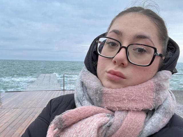 MayAlisa with glasses, pink and gray scarf, black puffy jacket, standing on wooden deck, ocean in background, cloudy sky, looking slightly to the side, pouty lips.