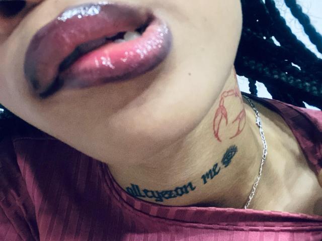 ElaHott's glossy pink lips are slightly parted, showing a hint of teeth. She has a tattoo below her jaw, a red outline tattoo, and wears a silver necklace.
