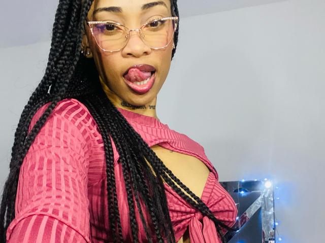 ElaHott with long black braids, pink glasses, and tongue out, wearing a pink ribbed top with a cutout, showing her tits. She's got a tattoo under her chin.