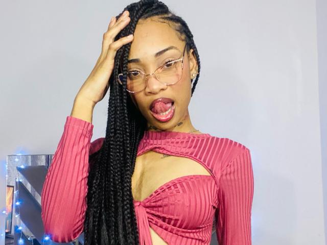 ElaHott com longas tranças pretas, blusa rosa texturizada que mostra os seios, óculos, língua de fora, mão no cabelo, com uma aparência fofa e sexy..