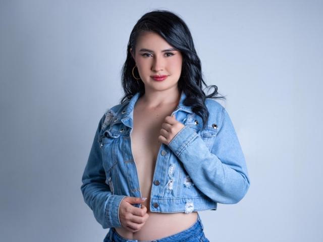 FrancheskaWaalsh with black hair, blue eyes, and pink lipstick, wearing a ripped blue denim jacket, open to show her tits, and blue jeans, standing against a light gray background.