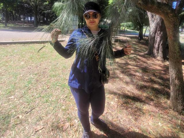 FrancheskaWaalsh with tits covered, wearing sunglasses, a dark blue jacket, black pants, and a black cap, holding pine needles, standing in a sunlit park.