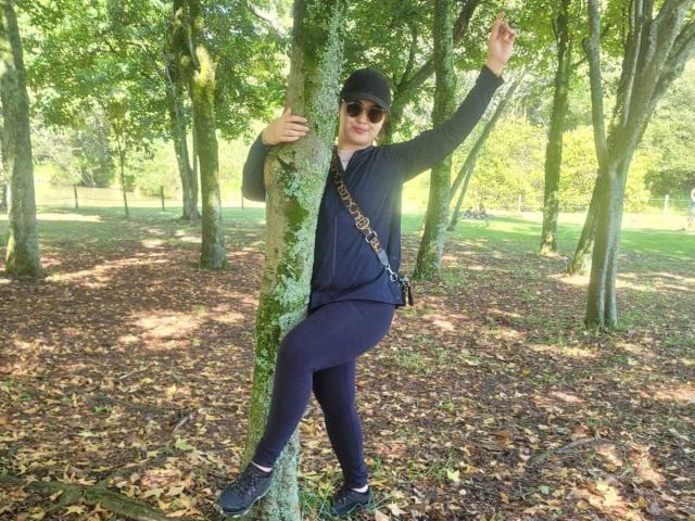 FrancheskaWaalsh, black cap, sunglasses, gray hoodie, black leggings, sneakers, sitting against a tree, right arm raised, left arm holding tree, leaf-covered ground, daytime, green trees.