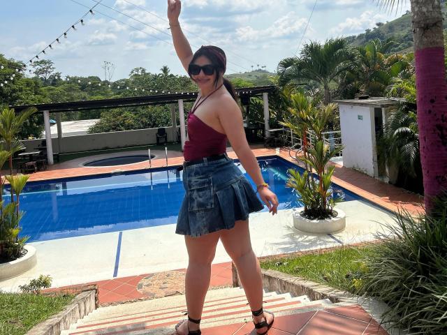 FrancheskaWaalsh stands by a blue pool, wearing a red strapless top, denim skirt, black heels, and sunglasses, with one arm raised, smiling, showing her ass.