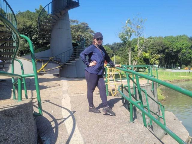 FrancheskaWaalsh with hands on green rail, wearing blue cap, sunglasses, navy long-sleeve shirt, black leggings, and black sneakers, standing on concrete by spiral staircase and water.