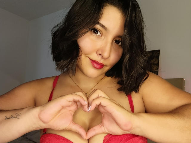Ketheryn with medium brown skin, black shoulder-length hair, red lipstick, and red lace bra, making a heart shape with her hands, showing her boobs. Tattoo on left arm.