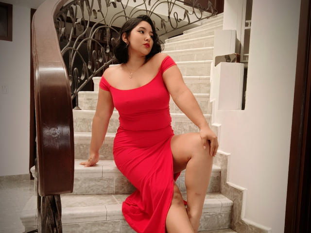 Ketheryn with curvy ass, sitting on marble stairs, wearing a red off-shoulder dress, one leg raised, dark wavy hair, red lipstick, wooden railing, ornate metal staircase.