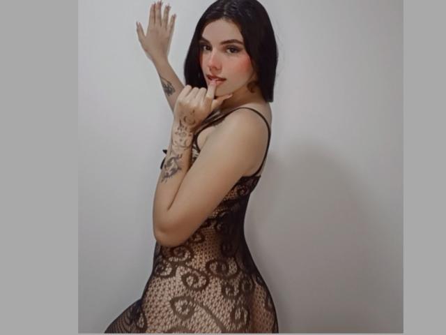GuadalupeClak mit dunklem Bob, trägt einen schwarzen Spitzen-Body, zeigt Tattoos an Händen und Armen, hat einen Finger im Mund, blickt zurück, die Hände an der Wand, wobei ihr Hintern und ihre Brüste sichtbar sind..