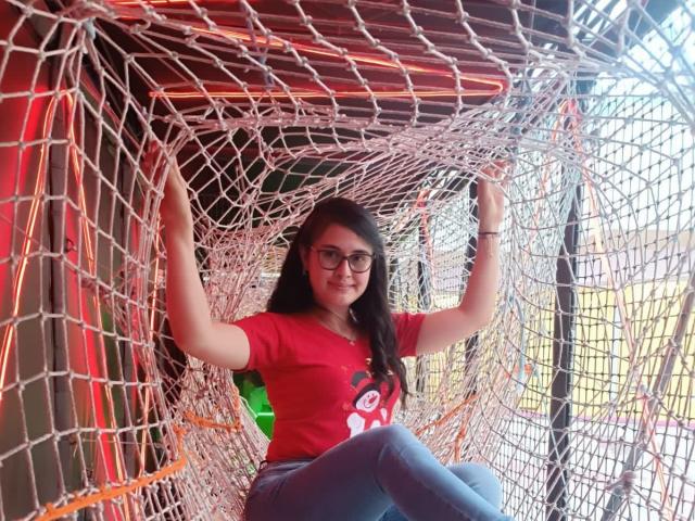 FrancheskaWaalsh with glasses, wearing a red shirt and blue jeans, sits in a white net structure, arms raised, smiling slightly, with long dark hair.