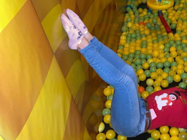 FrancheskaWaalsh's legs are up against a yellow wall, wearing blue jeans and pink socks, with a red shirt showing a white logo, in a ball pit.