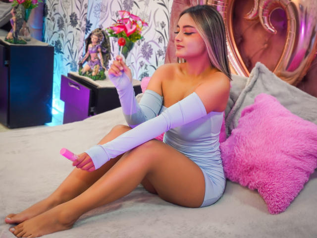 CloeesSuarez with long blonde hair, wearing a tight, off-shoulder, light blue dress, and white arm sleeves, sitting on a bed, putting on pink lipstick. Her ass is exposed.