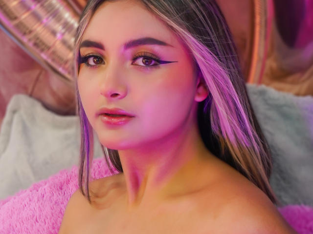 CloeesSuarez met roze en paarse verlichting, lang, recht bruin haar, opvallende eyeliner, roze lippenstift en onbedekte schouders, kijkt naar de camera met een lichte glimlach. Zachte, roze en grijze texturen..